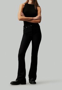 Jeans negros de corte acampanado y cintura alta, con cinco bolsillos y sutiles detalles de costura. Combinados con un top negro sin mangas y zapatos brillantes.