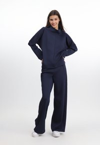 Trening de marină realizat dintr-un material neted, cu accente din dantelă texturată pe mâneci și laterale, având o croială lejeră și pantaloni evazați.