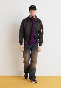 Black bomber jacket with ribbed cuffs, purple shirt, distressed blue jeans, and black sneakers. Cap with logo completes the outfit.