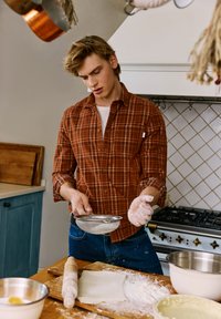 Un hombre con una camisa de cuadros marrón tamiza harina con un colador de metal. Un rodillo y recipientes para mezclar están sobre una encimera de madera de cocina. Hay polvo de harina visible.