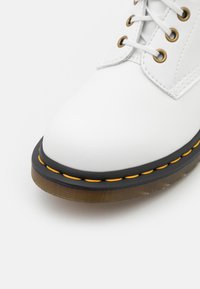 Bota de cuero blanca con punta redonda, cordones blancos y ojales dorados. Suela de goma negra con costuras amarillas y acento de tacón transparente.