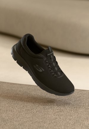 Sort Skechers sneaker med elastiske snørebånd, der svæver over en beige tekstureret overflade og neutral baggrund.
