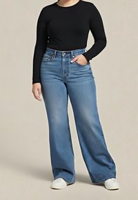 Personne portant un haut noir à manches longues, un jean bleu à jambes larges et des baskets blanches, debout avec une main dans la poche sur un fond uni.
