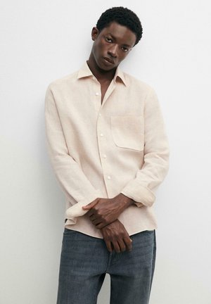 Giovane uomo con camicia beige a maniche lunghe abbottonata con taschino sul petto e jeans blu, in piedi contro un muro bianco uniforme, con le mani intrecciate.