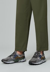 Piedi con sneakers multistrato in tonalità di grigio, verde e blu, abbinate a pantaloni a gamba larga verde oliva su uno sfondo neutro.