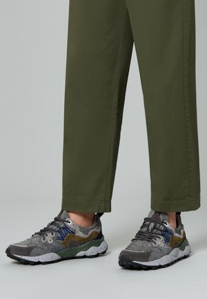 Piedi con sneakers multistrato in tonalità di grigio, verde e blu, abbinate a pantaloni a gamba larga verde oliva su uno sfondo neutro.