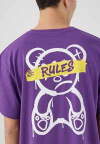 T-shirt in cotone viola con una grande grafica di un orso bianco e il testo "RULES" striato di giallo sopra gli occhi, con dettagli che gocciolano.