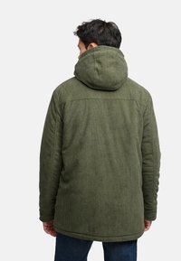 Olivgrüner Cordmantel mit großer Kapuze, der strukturierte vertikale Streifen und einen geraden Saum aufweist, getragen über blauen Jeans.