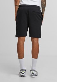 Man wearing black shorts, white socks, and white athletic shoes, standing with hands in pockets, showing tattooed arms.