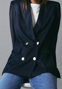 Persona che indossa un blazer doppiopetto blu navy con bottoni argento, una camicia bianca e jeans blu, seduta contro uno sfondo neutro.