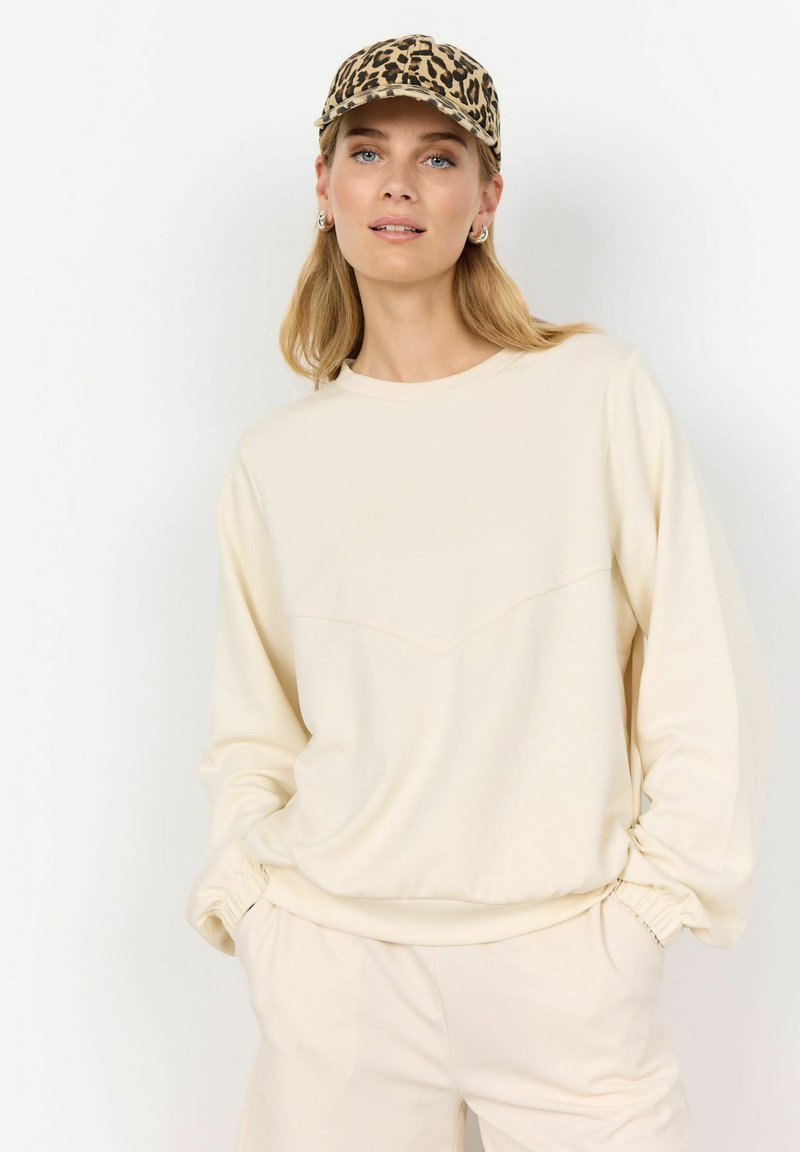 Cream-colored sweatshirt with a rounded neckline and elastic cuffs; features a subtle chevron seam detail across the chest.