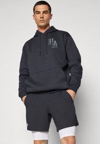 Mørkegrå hættetrøje med kengurulomme og snore, prydet med et fremtrædende UA-logo i en lidt lysere nuance. Et par matchende shorts.