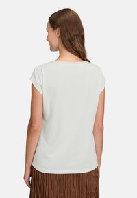 Biała koszulka z krótkim rękawem, okrągłym dekoltem, gładką fakturą i luźnym krojem, noszona z brązową spódnicą plisowaną.