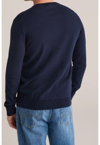 Tmavě modrý pletený svetr s kulatým výstřihem, žebrovanými manžetami a lemem, v kombinaci se světle modrými džínami. Hladká textura, minimální designové detaily.