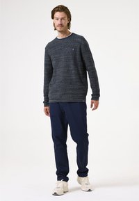 Navy gestreifter Pullover mit Rundhalsausschnitt, kombiniert mit navyfarbener Hose und cremefarbenen Sneakers. Verfügt über ein kleines Logo-Emblem auf der Brust.