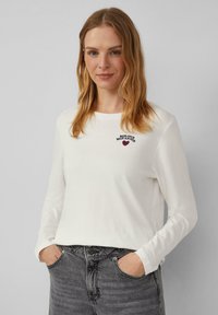 Witte lange mouwen shirt met ronde hals, voorzien van de tekst "BONJOUR MON AMOUR" en een hartborduursel. Gecombineerd met donkergrijze jeans.