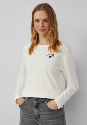 Chemise blanche à manches longues avec col rond, ornée du texte "BONJOUR MON AMOUR" et d'une broderie en forme de cœur. Associée à un jean gris foncé.