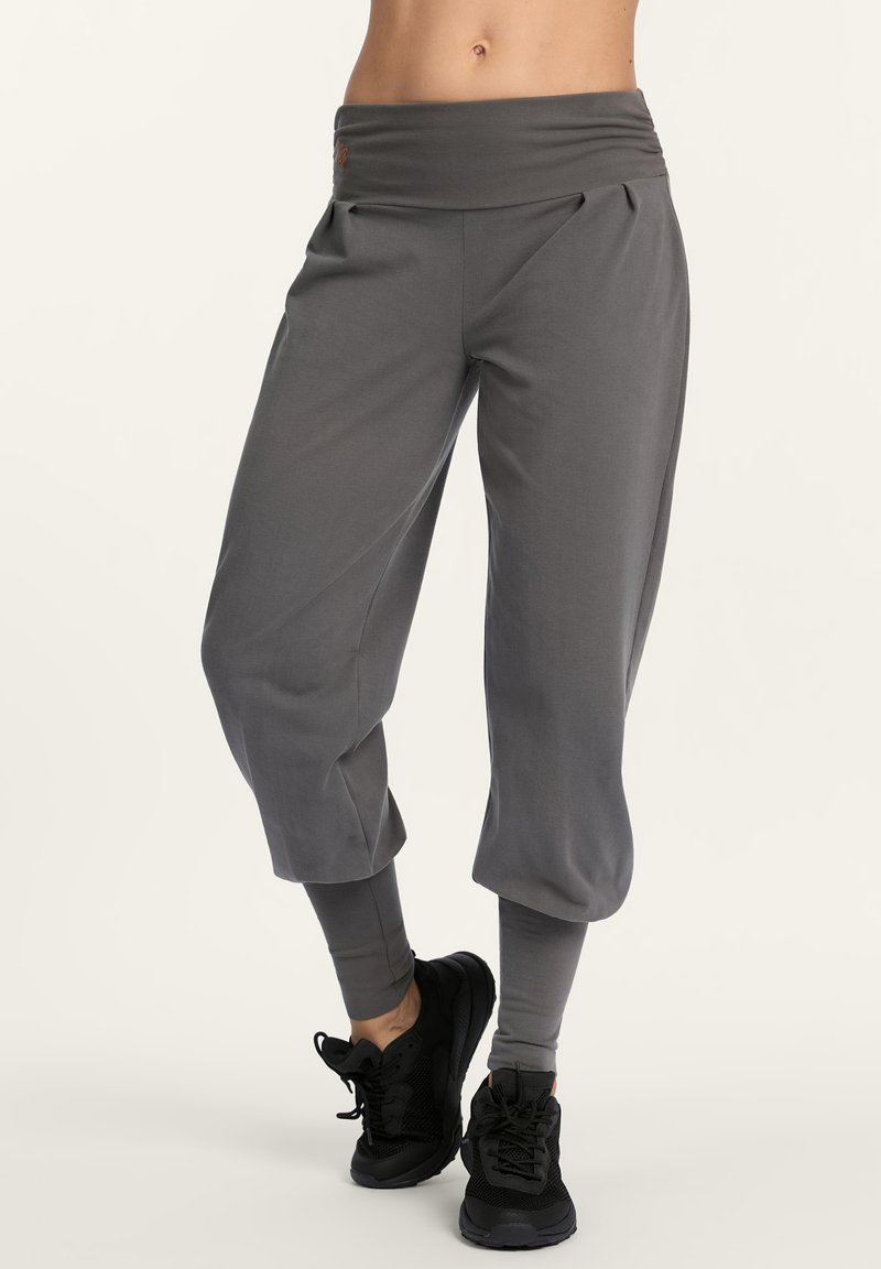 Person, der lockere graue, knöchellange Hosen mit eng anliegenden Knöcheln und schwarze Sportschuhe trägt, und vor einem einfarbigen hellen Hintergrund steht.