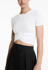 T-shirt bianco corto con maniche corte, dotato di un dettaglio arricciato frontale, realizzato in un tessuto liscio e elasticizzato. Indossato con pantaloni neri a vita alta.