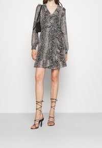 Frau trägt ein knielanges, langärmliges Wickelkleid aus Schlangenhaut mit Rüschen, schwarze Schnürsandalen mit hohen Absätzen und eine schwarze Schultertasche.