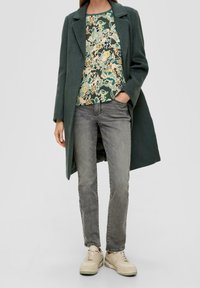 Dlouhý zelený kabát, vzorovaný krémově zelený top, šedé džíny, béžové tenisky. Měkké textury a vrstvený design.
