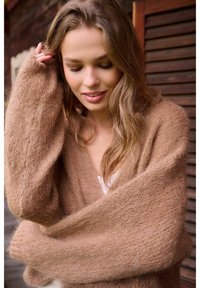 Mladá žena s dlhými vlnitými vlasmi, oblečená v mäkkom, veľkorysom hnedom svetri, dotýkajúca sa vlasov, pozerajúca sa dole s jemným úsmevom.