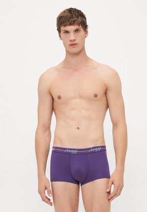 Junger Mann mit kurzem Haar, der lila Boxershorts mit "sloggi"-Logo am Bund trägt, vor einem schlichten weißen Hintergrund stehend.