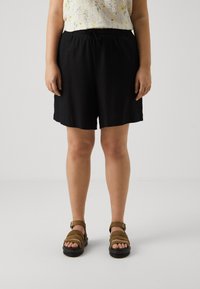 Shorts noirs en tissu doux, avec une taille élastique et un cordon de serrage. Associés à des sandales brunes à lanières.