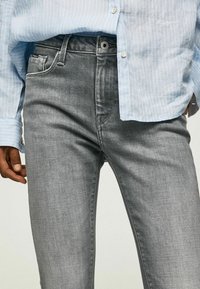 Jeans de mezclilla gris con un corte ajustado, que cuentan con bolsillos frontales, cierre de botón y una textura sutil. Combinados con una camisa a rayas de color azul claro.
