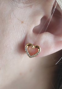 Herzförmige Ohrringe aus Gold mit ausgeschnittenem Zentrum, am Rand mit klaren Kristallen akzentuiert. Tragen Sie sie in einer Nahaufnahme eines Ohrs.