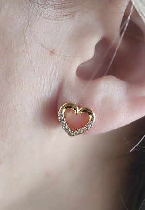 Orecchino a forma di cuore in oro, con un centro ritagliato, accentuato da cristalli trasparenti lungo il bordo. Indossato in un primo piano di un orecchio.