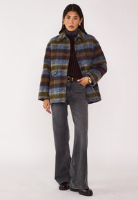Cappotto di lana a quadri con strisce blu, verdi e marroni; presenta un colletto, due tasche frontali e bottoni. Abbinato a jeans svasati grigio scuro.
