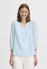 Blusa azzurro chiaro con scollo a V e maniche a tre quarti trasparenti. Presenta motivi a foglia in rilievo e una vestibilità comoda.
