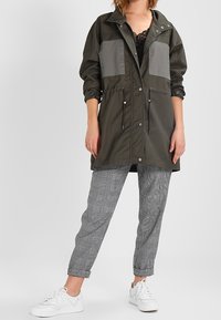 Kaki lättviktsjacka med grå fickor, dragsko i midjan och tryckknappar. Tillsammans med rutiga byxor och vita sneakers.