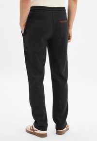 Black sweatpants with a smooth texture, elastic waistband, side pockets, and a small brown leather accent on the back pocket.