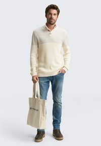 Crèmekleurige gebreide trui met geribbelde textuur, kraag met twee knopen; lichte denim jeans, bruine laarzen, een canvas totebag vasthoudend.