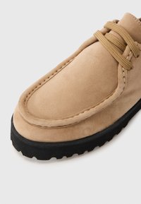Tan semsket sko med avrundet tå, sydd detaljer og brune snører; har en teksturert svart gummisåle.