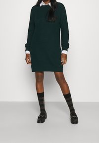 Abito lavorato a maglia di colore verde scuro con maniche lunghe, scollatura a girocollo e texture a coste. Abbinato a calze a quadri nere e rosse e mocassini neri.