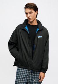 Jeune homme portant une veste noire à fermeture éclair avec une doublure bleue et un logo « HUGO », associée à un pantalon à carreaux bleus et gris, debout avec une main dans la poche.