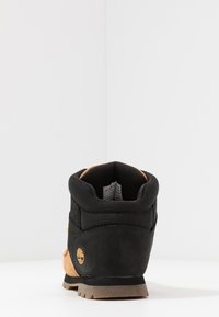 Las botas de senderismo impermeables en negro y marrón cuentan con un cuello acolchado, suela de goma y un logotipo amarillo. La parte superior combina cuero y malla transpirable.