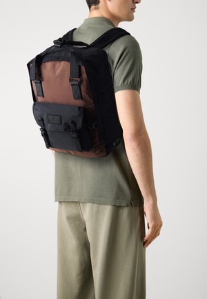 Sac à dos avec un design noir et marron, doté de bretelles ajustables, d'une poche avant et d'une finition texturée. Le matériau est un tissu robuste.