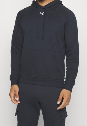 Mann trägt einen schwarzen Under Armour Hoodie mit Fronttasche und schwarze Cargo-Hose vor einfarbigem Hintergrund.