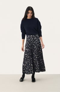 Un pull tricoté bleu marine associé à une jupe longue plissée ornée d'un imprimé floral dans des teintes crème et or, complété par des chaussures noires et des collants.
