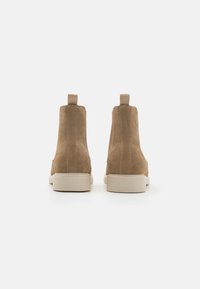 Botas de tobillo de ante color tostado con una textura suave, paneles laterales elásticos y una suela de goma de color claro. Cuenta con un lazo en el talón para facilitar su uso.