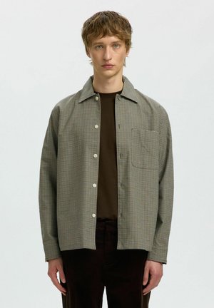 Jeune homme aux cheveux courts et ondulés portant une chemise à carreaux boutonnée sur une chemise marron foncé, sur un fond clair uni.