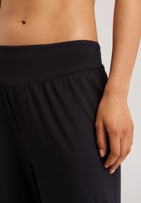 Czarne, miękkie spodnie dresowe z szerokim, elastycznym pasem i marszczeniami w okolicy talii. Zbliżenie na pas i ułożenie ręki.