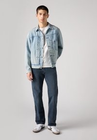 Jaqueta de denim azul claro com fecho de botão e dois bolsos, usada sobre uma t-shirt branca, combinada com jeans azul escuro e ténis brancos.
