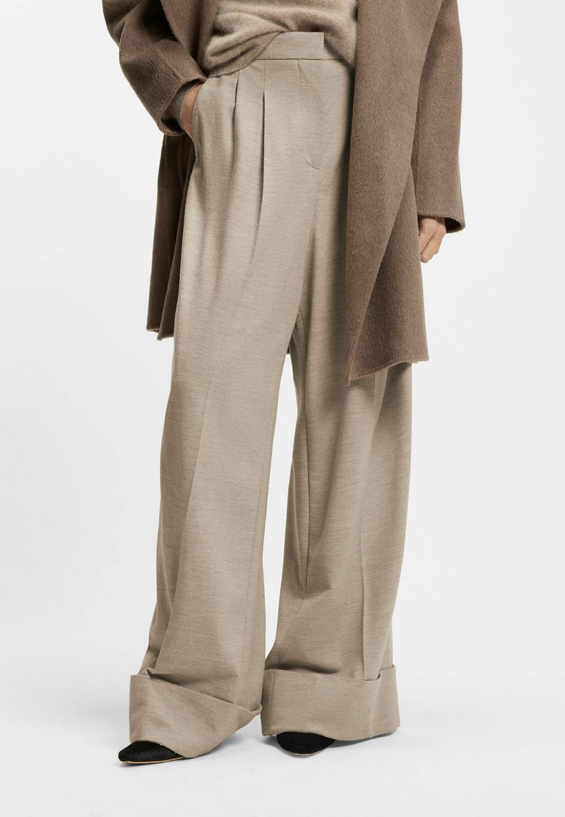 Beige wide-leg trousers with pleats, made of smooth fabric. Features a high waistband and rolled cuffs, paired with black pointed shoes.