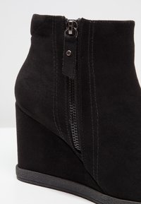 Botte de cheville à semelle compensée en daim noir avec un détail de fermeture éclair sur le côté, agrémentée de coutures et d'une semelle en caoutchouc texturée.