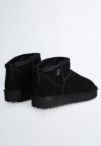 Botas de tobillo de ante negro con forro de piel sintética negra, suelas gruesas con estrías y pequeño logo metálico en el lateral, mostradas sobre un fondo blanco.